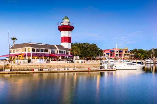 A scenic harbor view featuring a red and white lighthouse, waterfront buildings, and boats on calm water.
