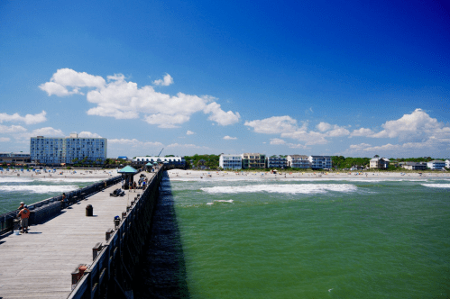 A wooden pier extends into a turquoise sea, with sandy beach and buildings under a bright blue sky with fluffy clouds.