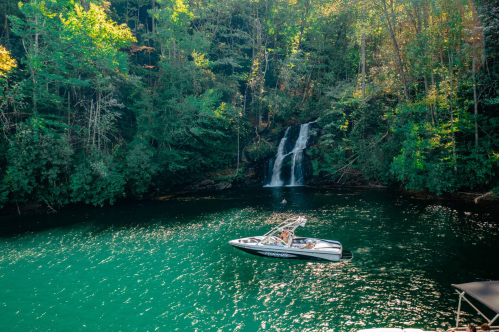 A boat floats on a serene green lake near a waterfall, surrounded by lush trees and vibrant nature.
