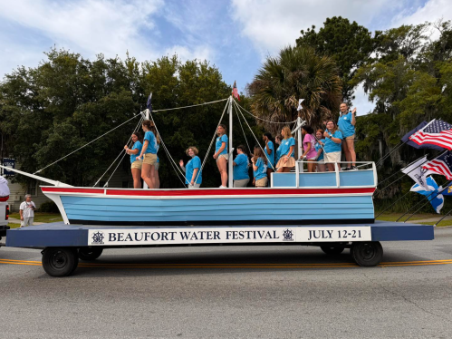 A colorful parade float shaped like a boat, featuring people in blue shirts, promoting the Beaufort Water Festival.