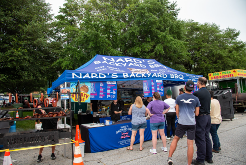 A food stall at an outdoor event with people waiting in line for BBQ, featuring a blue tent and menu boards.
