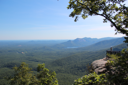 A panoramic view of lush green mountains and a lake under a clear blue sky, seen from a rocky overlook.
