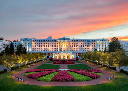 A grand hotel with a white facade, surrounded by colorful flower beds and trees, under a vibrant sunset sky.
