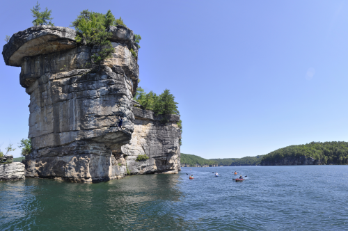 A panoramic view of a rocky cliff with trees, overlooking a calm lake and distant green hills under a clear blue sky.