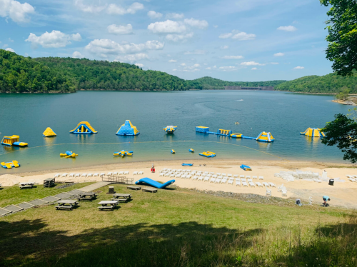 A scenic view of a lake with inflatable water attractions and a sandy beach, surrounded by green hills and blue skies.