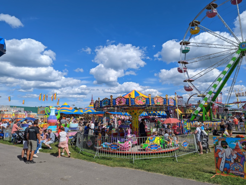 A vibrant fair scene with a Ferris wheel, carousel, colorful tents, and crowds enjoying various attractions under a blue sky.