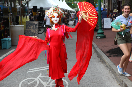 A performer in a vibrant red costume and fan poses on a street during a festival, with onlookers in the background.