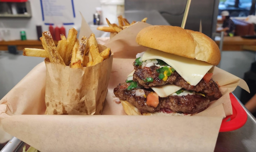 A stacked burger with cheese, onions, and tomatoes, served with a side of crispy fries in a paper container.