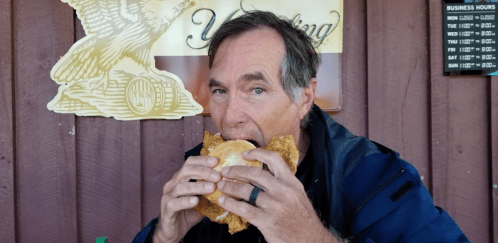 A man is about to take a big bite of a large fried chicken sandwich, sitting in front of a wooden wall.