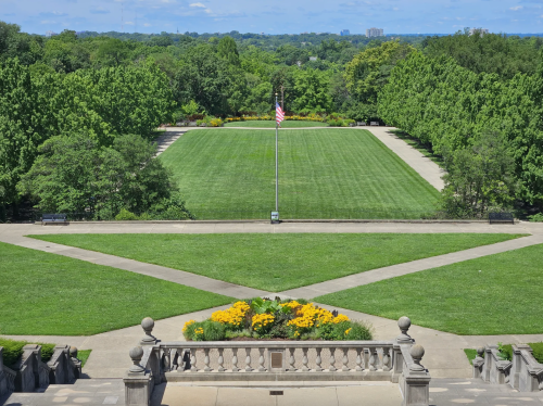 A wide view of a green park with neatly trimmed grass, flower beds, and an American flag in the background.