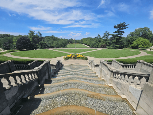 A scenic view of a landscaped garden with a fountain, steps, and lush greenery under a blue sky.