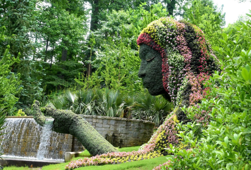 A large, floral sculpture of a woman surrounded by greenery and a waterfall in a lush garden setting.