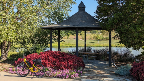 A gazebo surrounded by colorful flowers and greenery, with a bicycle parked nearby and a serene pond in the background.