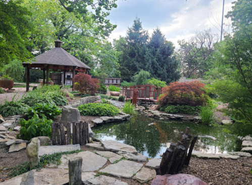 A serene garden scene featuring a gazebo, pond, bridge, and lush greenery under a cloudy sky.