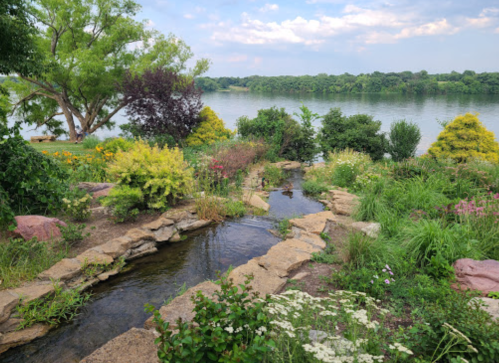 A serene garden by a river, featuring lush greenery, colorful flowers, and a small stream winding through the landscape.