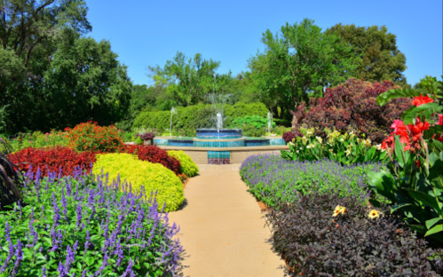 A vibrant garden path lined with colorful flowers leads to a fountain surrounded by lush greenery under a clear blue sky.