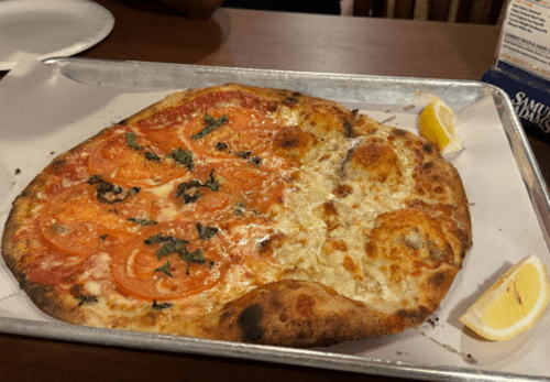 A half-and-half pizza on a metal tray, featuring fresh tomatoes and herbs on one side, and a cheesy topping on the other.