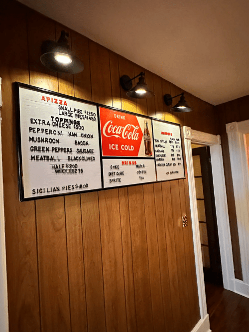 A vintage menu board on a wooden wall featuring pizza options and Coca-Cola advertisements.