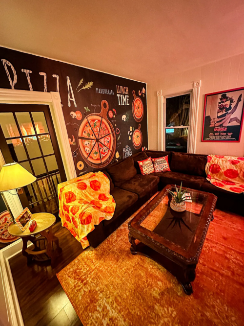 Cozy living room with pizza-themed wall art, a brown couch, and a warm ambiance from a lamp and window light.