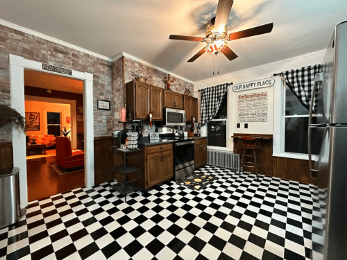 A cozy kitchen with wooden cabinets, black and white checkered floor, and a ceiling fan, featuring warm lighting and decor.