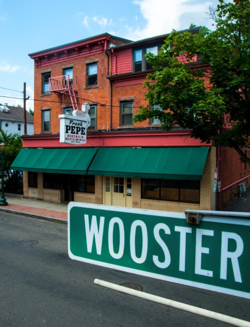 A brick building with green awnings houses Frank Pepe Pizzeria, with a street sign for Wooster in the foreground.