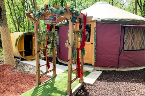 A wooden arch adorned with greenery leads to a cozy yurt surrounded by trees and a pathway.