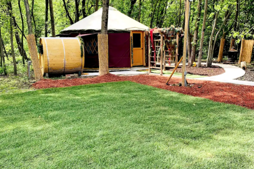 A grassy area with a yurt, wooden barrel, and playground equipment surrounded by trees and mulch.