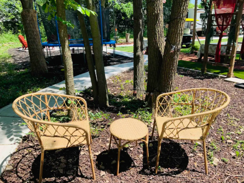 Two wicker chairs and a small table nestled between trees in a garden, with a trampoline visible in the background.