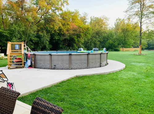 A round above-ground pool surrounded by green grass and trees, with a wooden deck and lounge chairs nearby.