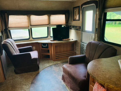 Cozy interior of a camper with two chairs, a TV, and large windows, featuring a circular rug and wooden accents.