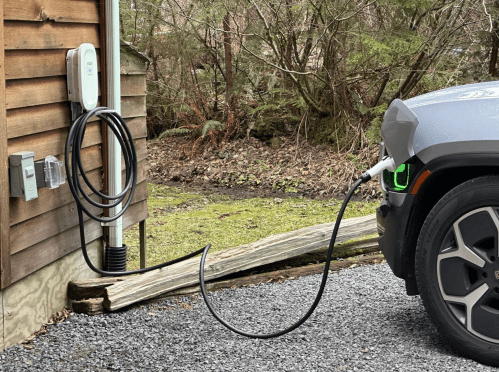 An electric vehicle is plugged into a charging station mounted on a wooden wall, with a green landscape in the background.