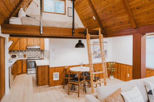 Cozy cabin interior with wooden beams, a lofted bed, kitchen, dining area, and a comfortable living space.