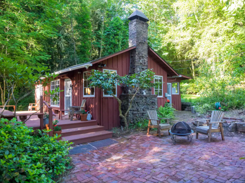 Cozy red cabin surrounded by lush greenery, featuring a stone chimney and a brick patio with outdoor seating.