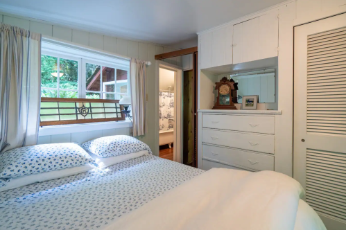 Cozy bedroom with a double bed, white linens, a dresser, and a window overlooking greenery.