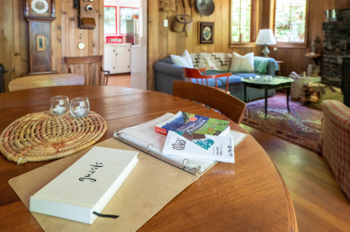 A cozy wooden living room with a dining table, binder, books, and a comfortable seating area in the background.