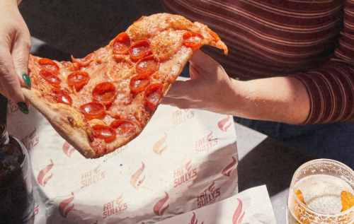A hand holds a large slice of pepperoni pizza above a table with drinks and branded paper.
