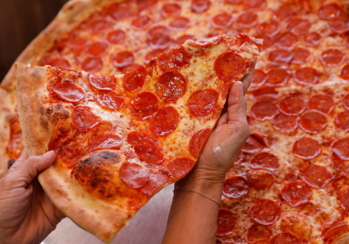 A hand holding a slice of pepperoni pizza, with a large pizza in the background.