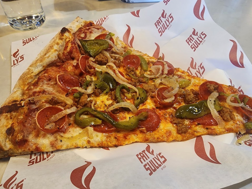 A slice of pizza topped with pepperoni, sausage, green peppers, and onions on branded paper.