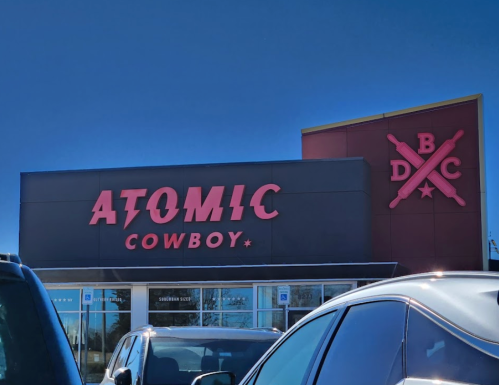 Sign for "Atomic Cowboy" with a blue sky in the background, viewed from a parking lot.