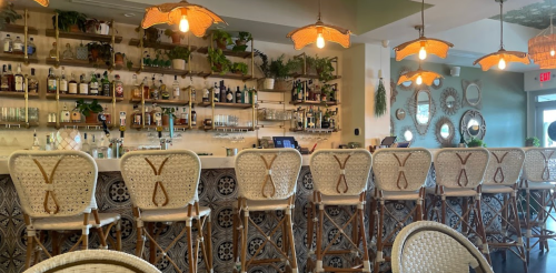 A stylish bar with rattan stools, shelves of liquor, and warm pendant lights in a modern, inviting space.
