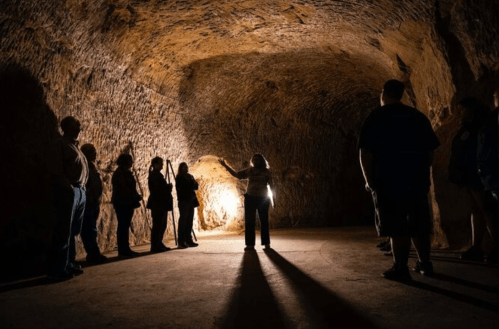 A group of people stands in a dimly lit cave, listening to a guide who gestures towards a light source.