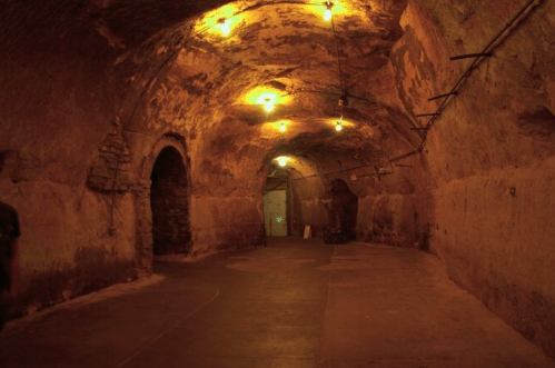 Dimly lit underground tunnel with arched ceilings and stone walls, featuring exposed light bulbs along the ceiling.