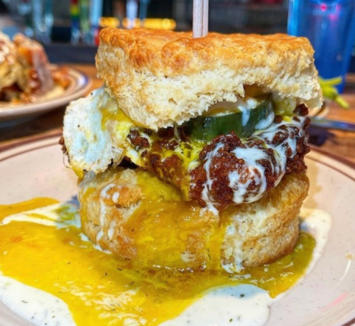 A stacked biscuit sandwich with fried chicken, pickles, cheese, and a drizzle of sauce, served on a plate.