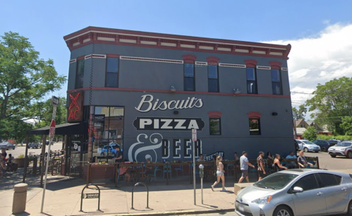 A gray building with a sign reading "Biscuits Pizza & Beer," featuring outdoor seating and pedestrians nearby.