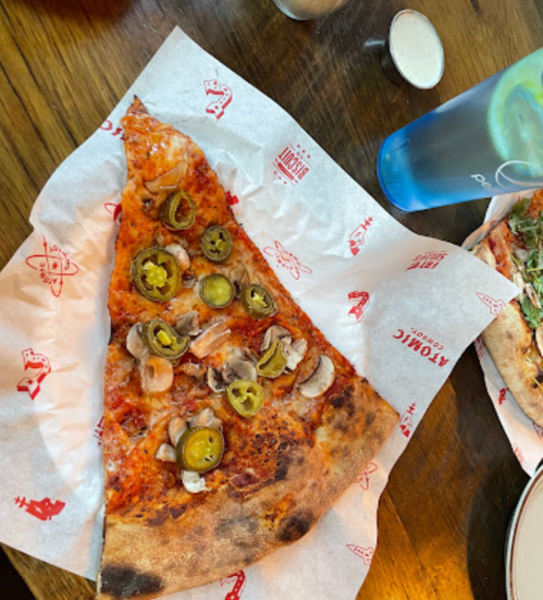 A slice of pizza topped with jalapeños and chicken, served on branded paper with a drink in the background.