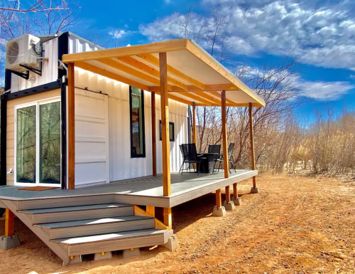 A modern container home with a wooden deck and patio, surrounded by sparse vegetation under a blue sky.