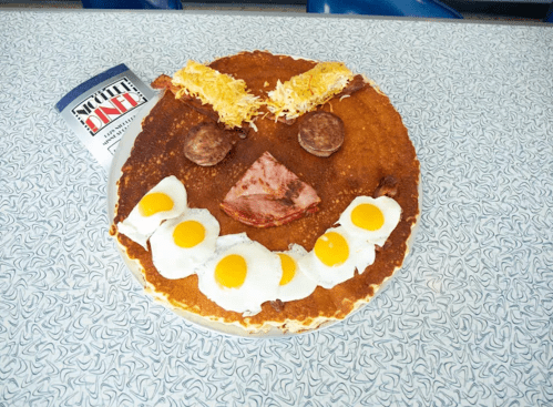 A large pancake decorated with bacon, sausage, cheese, and eggs to resemble a smiling face on a patterned table.