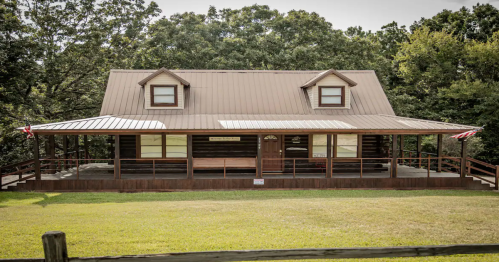 A large, rustic two-story cabin with a metal roof, surrounded by trees and a grassy lawn.