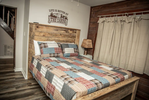 Cozy cabin bedroom with a wooden bed, plaid bedding, and a rustic lamp, featuring a sign that reads "Life is better at the cabin."