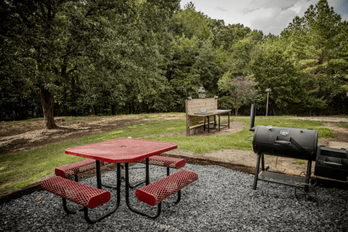 A red picnic table with benches near a grill and a stone cooking area, surrounded by trees and grass.